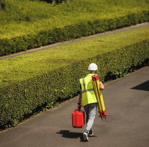 Landmeter met helm en fluohesje stapt met statief en Leica koffer langs een haag