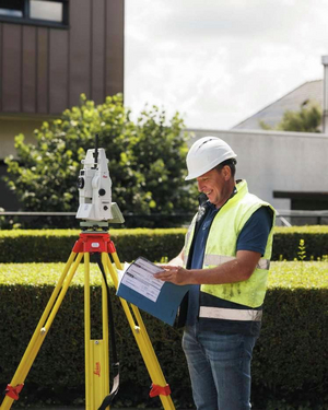 Landmeter met helm en fluohesje bekijkt een plan naast een Leica totaalstation