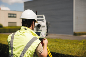 Landmeter met helm en fluohesje kijkt door een totaalstation op een bedrijfsterrein
