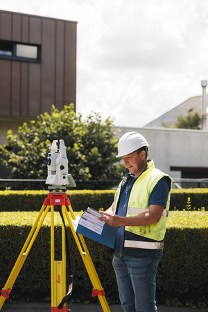Landmeter met helm en fluohesje bekijkt een plan naast een Leica totaalstation