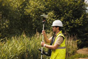 Landmeter met helm en fluohesje meet een punt op met een GNSS-meetstok in het groen