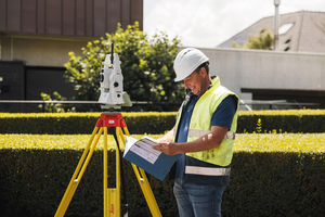 Landmeter met helm en fluohesje bekijkt een plan naast een Leica totaalstation