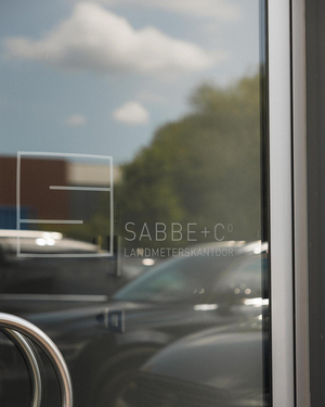 Logo van Sabbe landmeterskantoor op het raam met weerspiegeling van de parking